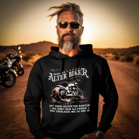 Ein Biker mit einem Hoodie für Motorradfahrer von Wingbikers mit dem Aufdruck, Alter Biker - Einige Kilometer runter, trinkt mehr - aber zuverlässig wie eh und je - in schwarz.
