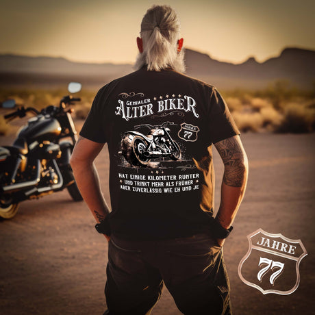 Ein Biker mit einem T-Shirt zum Geburtstag für Motorradfahrer von Wingbikers mit dem Aufdruck, Alter Biker - 77 Jahre - Einige Kilometer runter, trinkt mehr - aber zuverlässig wie eh und je - mit Back Print, in schwarz.