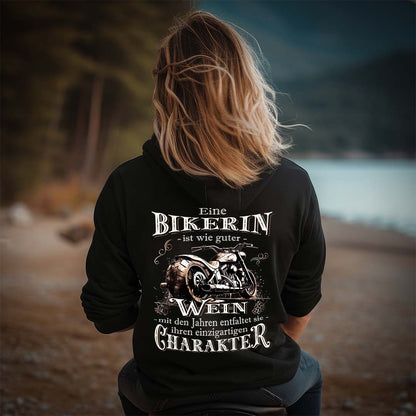 Eine Bikerin mit einem Hoodie für Motorradfahrerinnen von Wingbikers mit dem Aufdruck, Eine Bikerin ist wie guter Wein - mit den Jahren entfaltet sie ihren einzigartigen Charakter - mit Back Print, in schwarz.