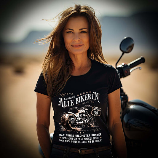 Eine Bikerin mit einem Geburtstags-T-Shirt für Motorradfahrerinnen von Wingbikers mit dem Aufdruck, Alte Bikerin - 50 Jahre - Einige Kilometer Runter - Doch elegant wie eh und je - in schwarz.