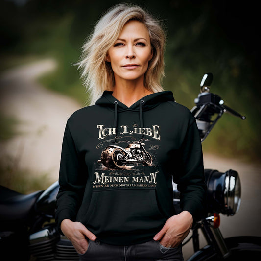 Eine Motorradfahrerin mit einem Bikerin Hoodie für Motorradfahrerinnen von Wingbikers mit dem Aufdruck, Ich liebe meinen Mann, wenn er mich Motorrad fahren lässt! - in schwarz.